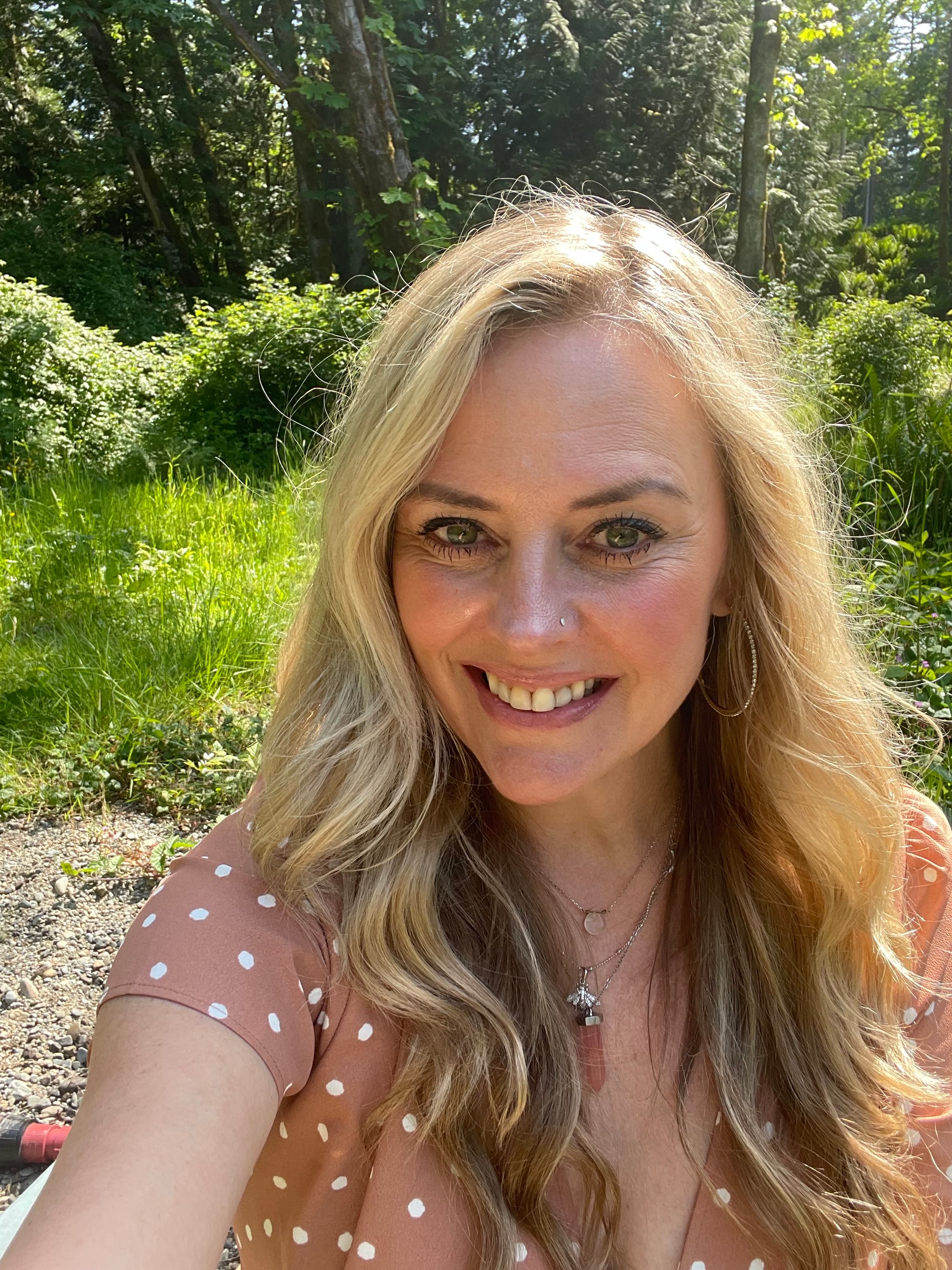 Smiling blonde woman with green eyes wearing a polka dot top in bright sunlight.